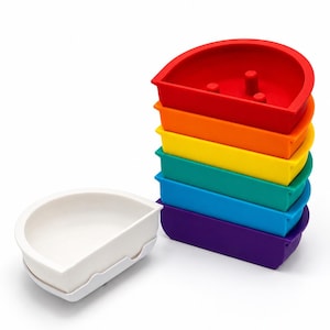 Puede incluir: Un conjunto de cuencos apilables semicirculares en una secuencia de colores arcoíris que incluye rojo, naranja, amarillo, verde, azul y morado, acompañado de un cuenco blanco de la misma forma. Cada cuenco tiene un borde plano y un lado curvo.