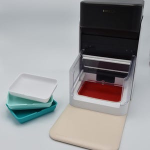 May include: A black and white appliance with a clear compartment and a red tray. Three square trays in white, teal, and turquoise are stacked to the left. The appliance sits on a beige mat.