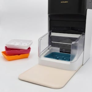 Puede incluir: Un comedero automático para mascotas con parte superior negra y base blanca. El comedero tiene un recipiente transparente y una alfombrilla beige. Tres bandejas de comida coloridas en blanco, rosa y naranja están apiladas a la izquierda. El texto "PETLIBRO" es visible.