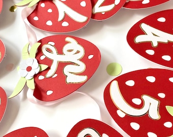 May include: A red and white strawberry-shaped banner with the numbers 1 through 12 printed in gold on each piece. The banner is decorated with white flowers and green leaves.