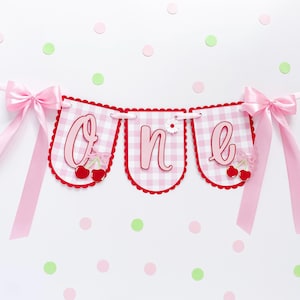 May include: A pink and white gingham banner spelling "ONE" in pink script. The banner is decorated with red cherries, bows, and a daisy. Pink ribbons and bows are attached to the banner's ends. The background is white with pink and green confetti.