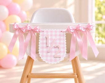 May include: A high chair banner featuring a scalloped-edge pennant with a light pink and white gingham pattern and the number 1/2 in raised lettering. The banner is accented with four light pink satin ribbon bows tied on each side.