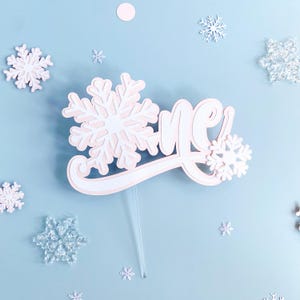 May include: White and pink cake topper with the word "One" and snowflake designs. The topper is on a clear plastic stick.