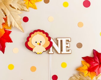 May include: A Thanksgiving-themed cake topper with a cartoon turkey and the word "ONE" in white letters. The turkey is red, orange, and brown with a white body and a red beak. The background is a light beige with scattered red, orange, and yellow dots.