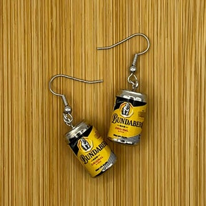 May include: A pair of silver earrings featuring miniature cans of Bundaberg Rum and Cola. The cans are yellow with black lettering and a white bear logo.