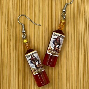 May include: A pair of earrings featuring miniature bottles of Captain Morgan's Spiced Gold rum. The bottles are made of clear plastic with a red liquid inside and a gold-coloured cap. The bottles have a label with the Captain Morgan logo and the words "Original Spiced Gold".