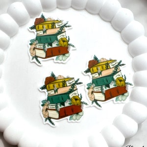 May include: Three stackable stickers featuring a pile of books with floral accents. The books are illustrated in shades of green, yellow, brown, and red.