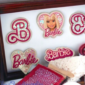 May include: A set of five pink and white glitter-covered iron-on patches featuring the word "Barbie" in a variety of styles, including a heart shape and a cursive font. The patches are displayed on a white background in a wooden frame.