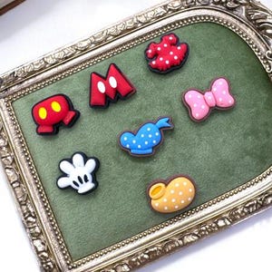 May include: A collection of colorful cartoon-themed charms displayed on a green velvet surface within an ornate, gold-framed tray. The charms include a red bow, a white glove, and other recognizable shapes. The Cloud 9 Supplies logo is visible.