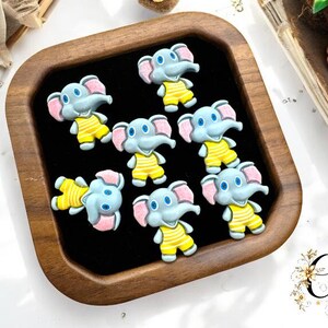 May include: A wooden tray with a black interior holds several small, cartoon elephant charms. The elephants are gray with pink ears and wear yellow overalls. The tray is surrounded by decorative items, including a succulent and a pinecone.