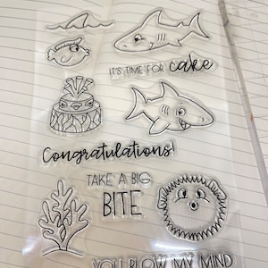 May include: Clear stamps with ocean-themed illustrations and phrases, including a shark, a fish, a cake, seaweed, a sea urchin, and the text "It's time for cake", "Congratulations!", "Take a big bite", and "You blow my mind".