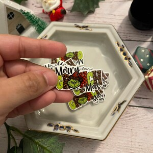 May include: A collection of Grinch-themed Christmas stickers featuring the Grinch's face and the text "Merry Grinchmas". The stickers are printed on a white background with a green, brown, and red color scheme.