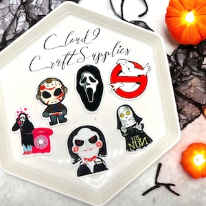 May include: A white octagonal plate with the text "Cloud 9 Craft Supplies" on it. The plate contains seven horror movie character iron-on patches. The patches are of Jason Voorhees, Ghostface, the Ghostbusters logo, the Nun, Jigsaw, and a character from the movie Scream.