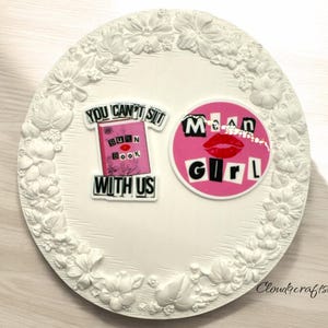 May include: White ceramic plate with a floral design border. Two pink and white stickers are on the plate. One sticker says "You can't sit with us" with a book that says "Burn Book" on it. The other sticker says "Mean Girl" with a red lip print.