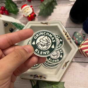 May include: A close-up of a green and white round sticker with the text "Grinchmas Blend" and a cartoon image of the Grinch. The sticker is being held by a hand.