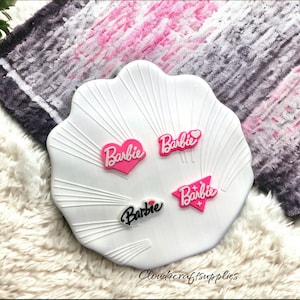 May include: Four pink and white plastic charms with the word "Barbie" in white lettering. The charms are in the shape of a heart, a star, and two different styles of a banner.