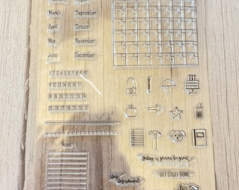 May include: Clear acrylic stamp set with a calendar, monthly planner, and various icons and phrases. The set includes a calendar grid, monthly planner, notes section, and icons such as a camera, pencil, umbrella, cake, apple, arrow, star, phone, notebook, hammer, dog, and a book. The set also includes phrases like "Stay Determined", "Goals", "Plans", "Today is gonna be great", and "Get Stuff Done".