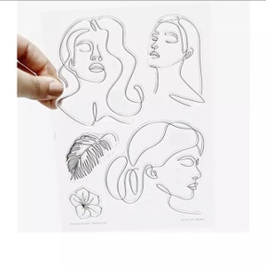 May include: A clear sheet of transparent stamps featuring four line drawings of faces and two line drawings of plants. The faces are of women with long hair and closed eyes. The plants are a feather and a flower.