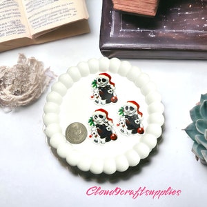 May include: A white, scalloped-edge plate holds three Jack Skellington-themed charms wearing Santa hats. A coin sits next to the charms. The charms are black, white, and red. A succulent plant and an open book are in the background.