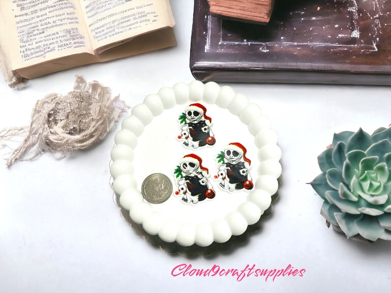 May include: A white, scalloped-edge plate holds three Jack Skellington-themed charms wearing Santa hats. A coin sits next to the charms. The charms are black, white, and red. A succulent plant and an open book are in the background.