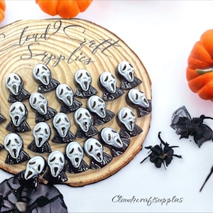 May include: A wooden round slice with a variety of small, silver and black, screaming face Halloween charms. The charms are arranged in a circular pattern. The background is white with two orange pumpkins and two black spiders.