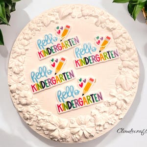 May include: Five colorful "Hello Kindergarten"  stickers with a pencil and hearts design. The stickers are on a white background with a floral design.
