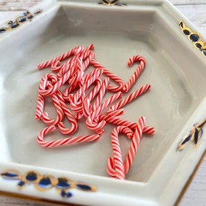 May include: A white ceramic dish with a gold and blue trim holds a pile of red and white candy canes.