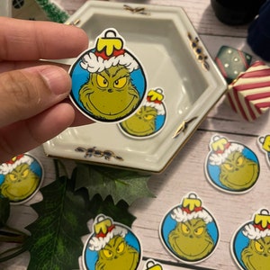 May include: A set of Grinch-themed Christmas ornaments. Each ornament features a cartoon image of the Grinch wearing a Santa hat and smiling. The ornaments are round and have a blue background.