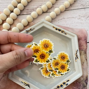 May include: A collection of sunflower stickers with yellow petals and green leaves, held near a light gray, hexagonal dish with gold and blue accents. A string of cream-colored wooden beads is in the background.
