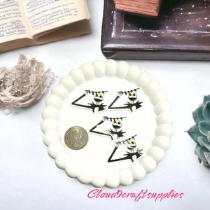 May include: A white, scalloped-edge plate holds several Jack Skellington-themed charms with colourful string lights. A silver coin sits near the charms. The background includes a succulent, an open book, and a wooden box.
