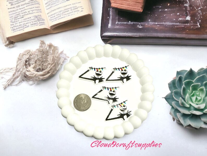 May include: A white, scalloped-edge plate holds several Jack Skellington-themed charms with colourful string lights. A silver coin sits near the charms. The background includes a succulent, an open book, and a wooden box.