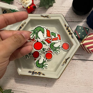 May include: A white hexagonal ceramic dish with a gold rim and blue and gold accents. The dish contains several small Christmas-themed stickers featuring a green Grinch-like figure with a red and white Santa hat and red ornaments.