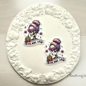 May include: Two colorful resin cabochons with a skull wearing sunglasses and holding a drink. The text "Cool Mums Club" is on a banner below each skull.