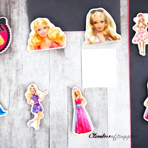 May include: A collection of Barbie doll cutouts in various poses and outfits. The cutouts feature blonde hair and different dresses, including a pink and white striped dress, a blue gown, and a purple dress. Some cutouts are in oval frames.