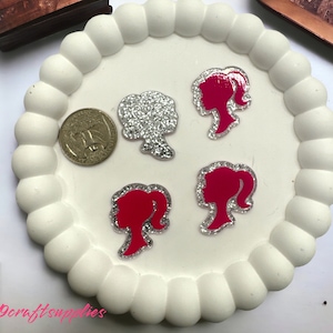 May include: Four decorative silhouette stickers of a woman's face with a ponytail. Two stickers are silver with a glittery finish and two stickers are pink with a glittery finish. The stickers are on a white plate with a textured edge.