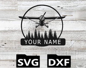 Op de afbeelding: Zwart metalen vliegtuigontwerp met een propeller, geplaatst in een cirkel boven een rij dennenbomen. De tekst "YOUR NAME" staat onder de bomen. Daaronder de letters "SVG" en "DXF" in aparte zwarte vakken.
