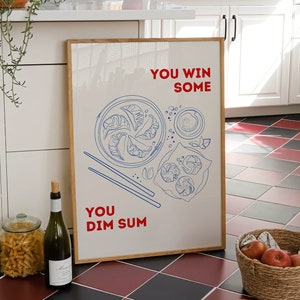 Op de afbeelding: Een ingelijst afdruk met een witte achtergrond en blauwe en rode tekst. De tekst luidt "You win some You Dim Sum". De afdruk toont een lijntekening van een bord met dim sum met eetstokjes.