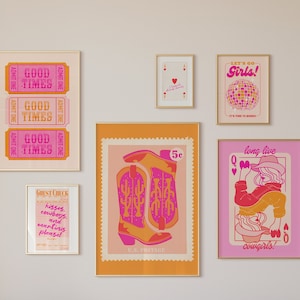 May include: A pink sofa with yellow and pink pillows in a living room with a white flower arrangement on a white coffee table. The wall behind the sofa is decorated with a collection of framed prints with a western theme, including a cowboy boot, a queen of hearts playing card, and a sign that says "Good Times".