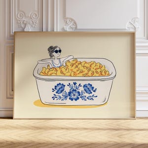 May include: A framed art print featuring a whimsical illustration of a person relaxing in a tub filled with macaroni and cheese. The tub is decorated with blue floral patterns, and the background is a light yellow.