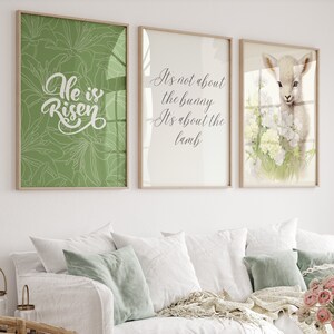 May include: Three framed prints with Easter themes. The first print has a green background with white floral designs and the text "He is Risen". The second print has a white background with the text "It's not about the bunny, it's about the lamb". The third print has a white background with a watercolor illustration of a white lamb with pink flowers.