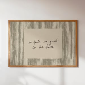 Può includere: Opera d'arte da parete con cornice in legno con la frase "it feels so good to be home" in grafia nera. Lo sfondo presenta un rettangolo beige con linee verticali grigie, creando un'estetica minimalista e neutra.