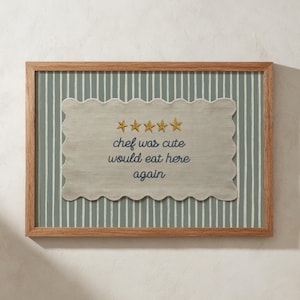 Puede incluir: Un cuadro enmarcado con un marco de madera marrón claro. La obra de arte presenta un diseño gris claro con bordes festoneados con el texto "chef was cute would eat here again" y cinco estrellas doradas, sobre un fondo a rayas.