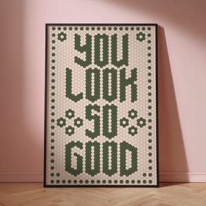 May include: Framed art print with the words "YOU LOOK SO GOOD" in green hexagonal tiles on a cream background. The artwork is framed in black and is displayed against a pink wall and wooden floor.