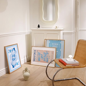 May include: A white room with a white chair, a white cabinet, and three framed posters. The posters are blue and pink with text that reads "GUEST CHECK", "YOU'RE LIKE REALLY PRETTY!" and "ADMIT ONE *GOOD TIMES*" The chair is a bentwood chair with a woven seat and back. The cabinet has a white oval mirror above it.