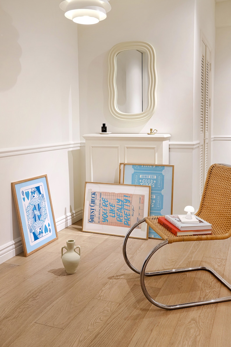 May include: A white room with a white chair, a white cabinet, and three framed posters. The posters are blue and pink with text that reads "GUEST CHECK", "YOU'RE LIKE REALLY PRETTY!" and "ADMIT ONE *GOOD TIMES*" The chair is a bentwood chair with a woven seat and back. The cabinet has a white oval mirror above it.