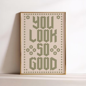 May include: A framed art print with the words "YOU LOOK SO GOOD" in a green, hexagonal tile design. The background is a light beige color, and the frame is gold. The artwork is displayed on a white shelf.
