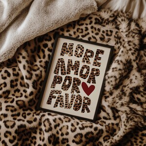 May include: A framed print with the text "More Amor Por Favor" in a leopard print font with a red heart in the middle of the text. The print is on a leopard print throw blanket.