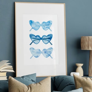 May include: A framed watercolor print of three pairs of blue heart-shaped sunglasses. The sunglasses are arranged vertically in a row.