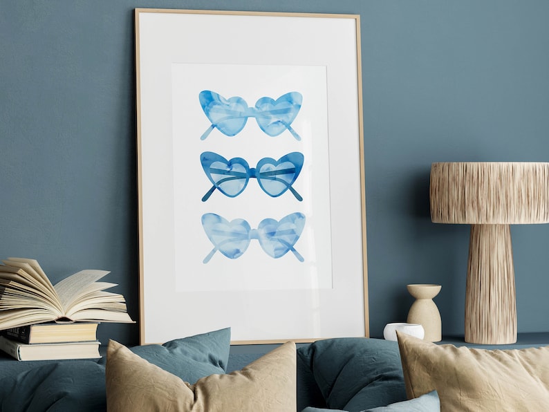 May include: A framed watercolor print of three pairs of blue heart-shaped sunglasses. The sunglasses are arranged vertically in a row.