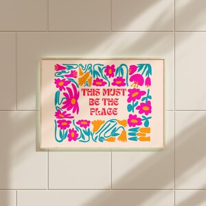 以下が含まれることがあります： ピンクとターコイズブルーの花柄プリントに、赤い文字で「This must be the place」と書かれています。プリントは明るい木製のフレームで額装されています。